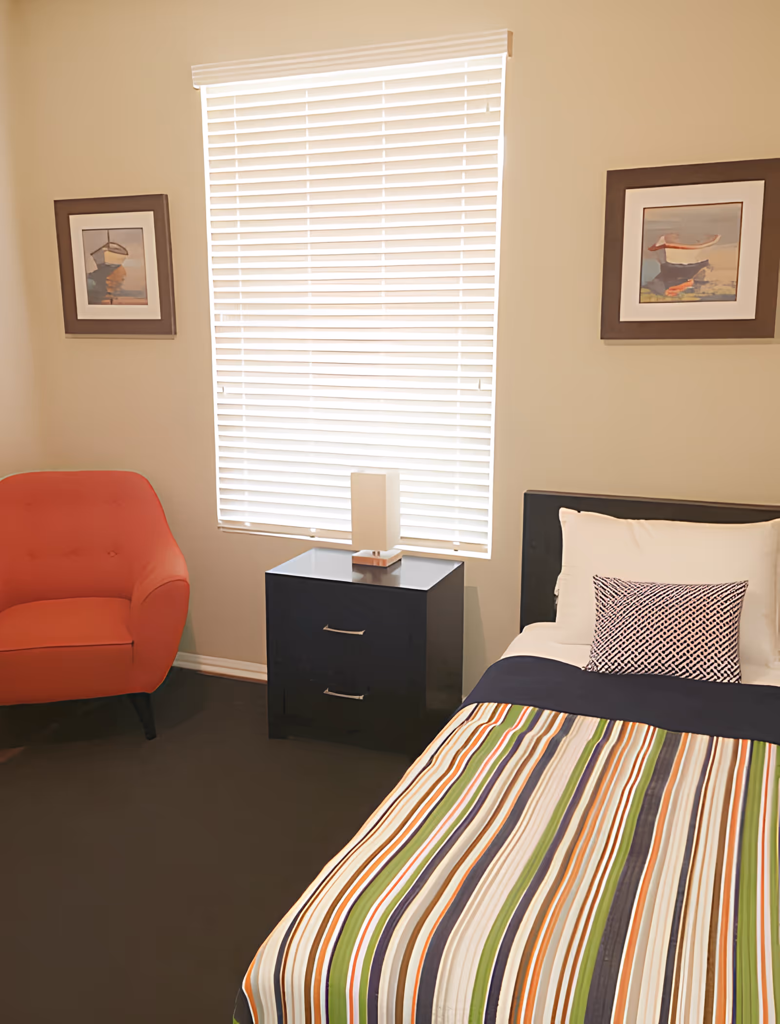 A bright single bedroom with a striped bedspread, black nightstand and lamp, a red armchair, and framed artwork beside a window with blinds.
