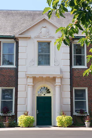 Front entrance of a brick building with a green door numbered 650, framed by decorative columns and potted plants.