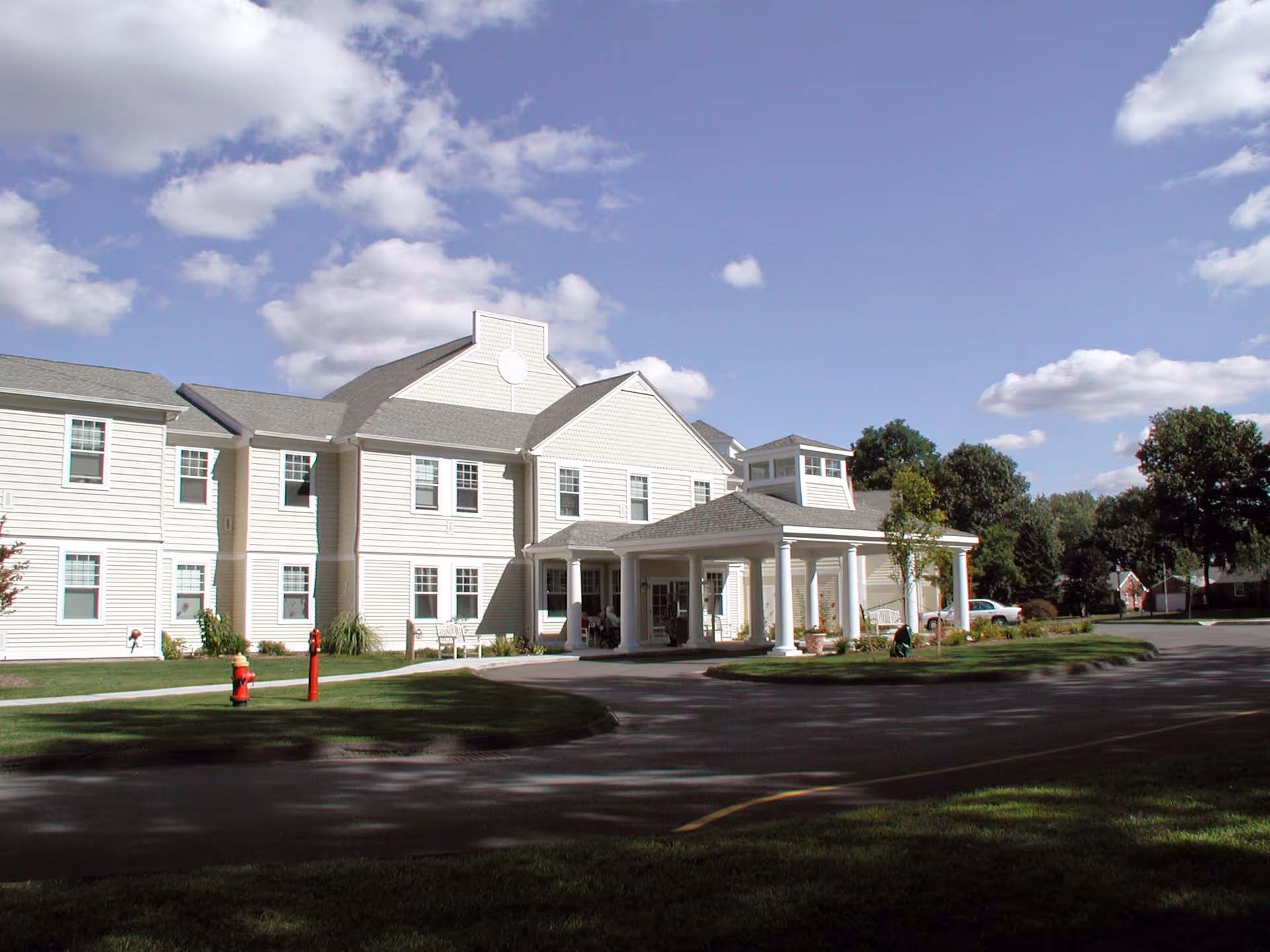 Front exterior of a two-story light-colored senior living building with a covered porte-cochère and driveway under a partly cloudy sky.