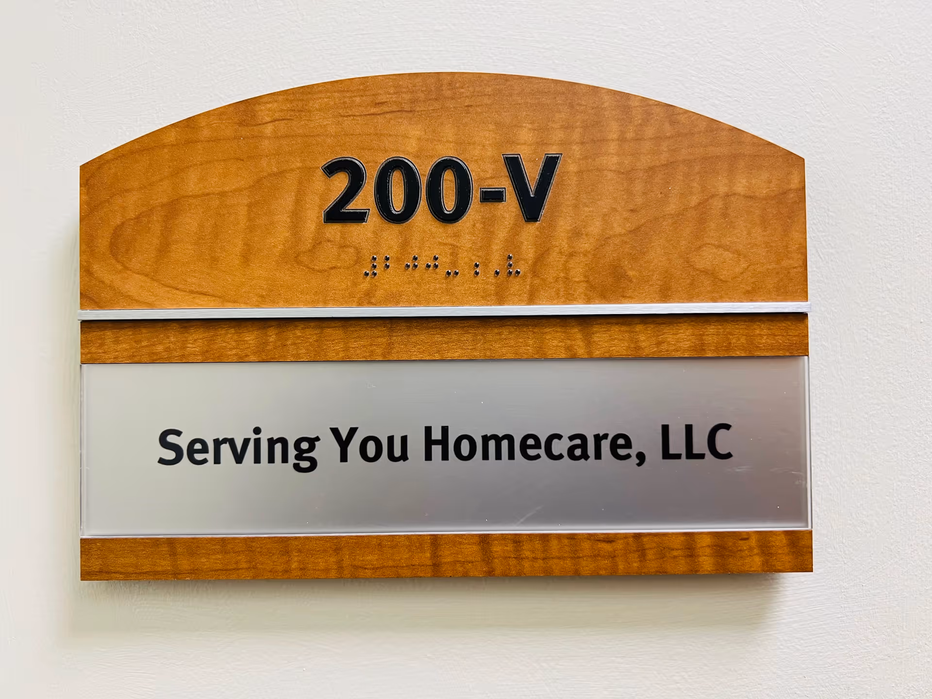 Wooden wall plaque with 200-V and Serving You Homecare, LLC on beige wall.