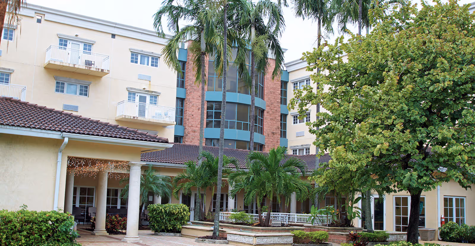 Exterior courtyard and front facade of a multi-story senior living building with palm trees, balconies, and covered walkways.