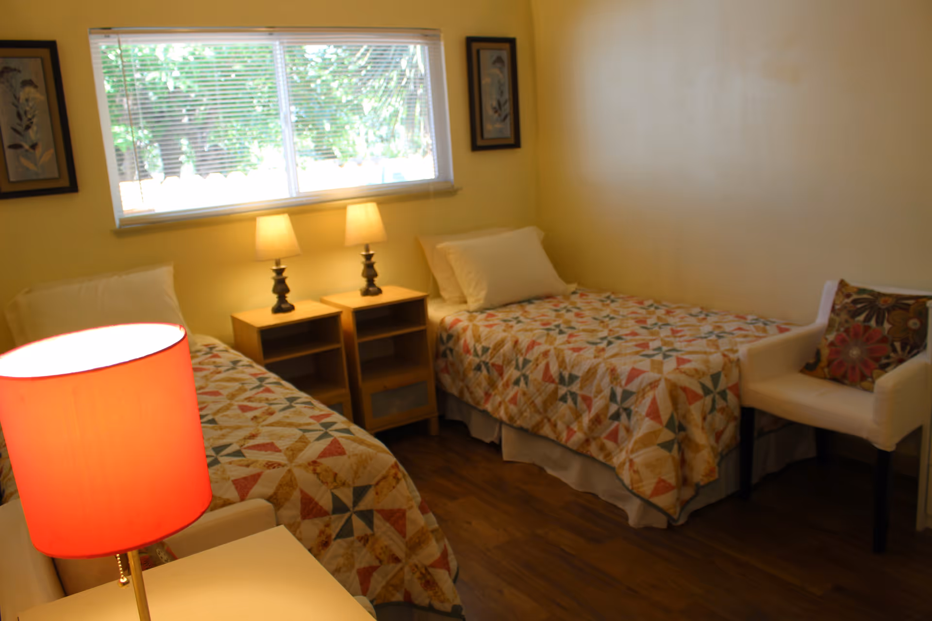 A cozy bedroom with two single beds covered in colorful patterned quilts. Between the beds are two wooden nightstands with matching lamps. A window with blinds lets in natural light, and there are framed pictures on the wall. A white chair with a floral cushion is positioned near one of the beds. The room has wooden flooring and light yellow walls.
