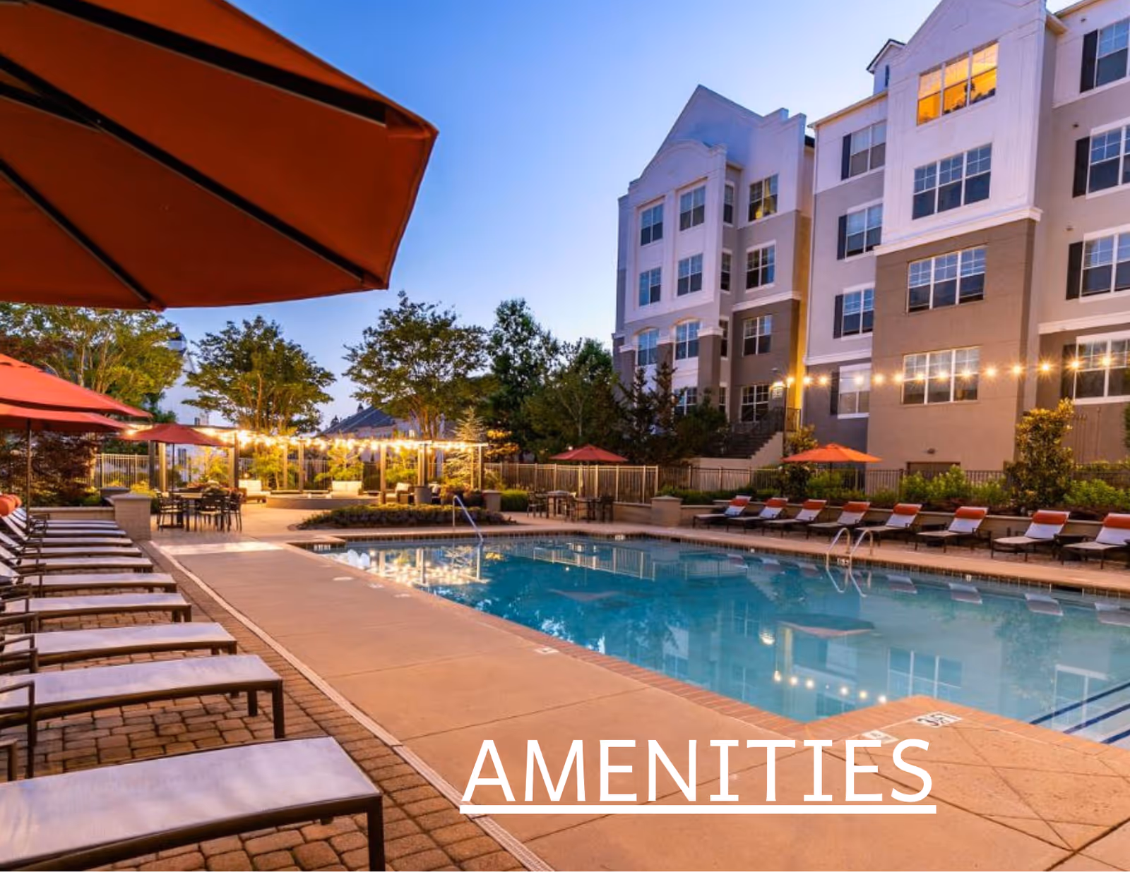 Outdoor swimming pool area with lounge chairs, red umbrellas, string lights, and a multi-story building in the background.