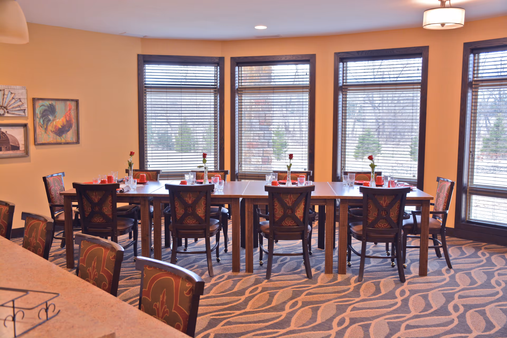 A dining room with a long rectangular table set for eight people. Each place setting includes a red napkin, glassware, and a single red rose in a vase. The room has large windows with blinds, patterned carpet, and warm yellow walls decorated with framed artwork.