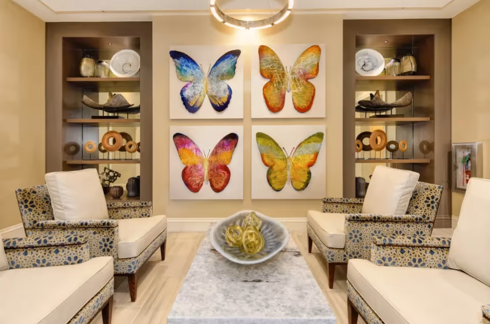 A cozy sitting area with four cushioned armchairs featuring patterned upholstery arranged around a rectangular marble coffee table. On the table is a decorative bowl with yellow glass spheres. The wall behind the chairs displays four colorful butterfly artworks in a 2x2 grid. Built-in shelves on either side of the artwork hold various decorative items. The room is warmly lit with a modern circular ceiling light.