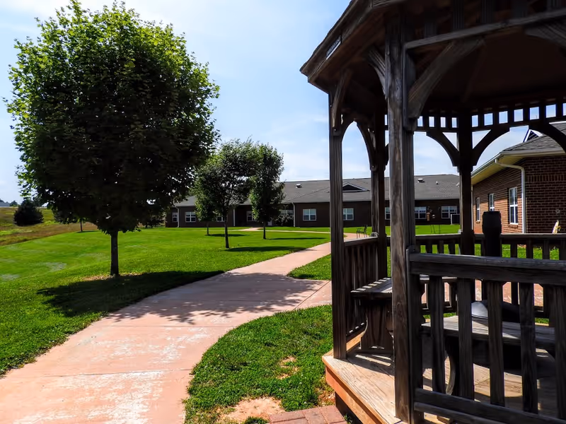 Outdoor view of a nursing home facility with a paved walkway, green lawn, several trees, and a wooden gazebo on the right side. The building is visible in the background under a clear blue sky.