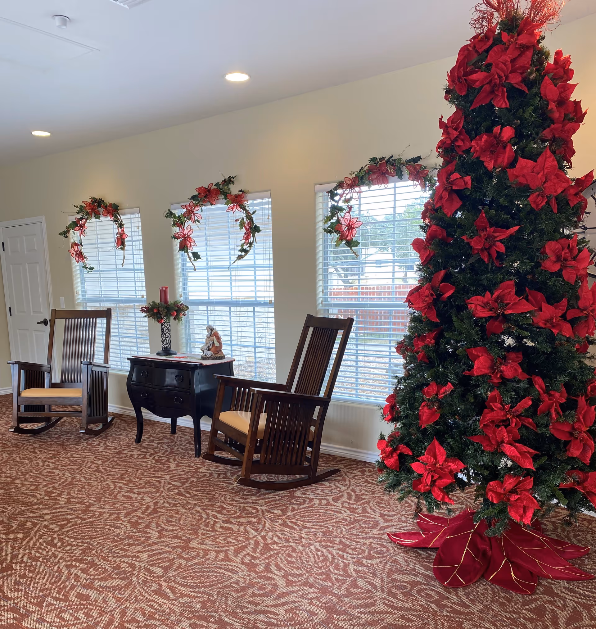 A decorated indoor seating area with two wooden rocking chairs and a small black table between them. The table holds a candle and a figurine. Three windows with white blinds are adorned with red and green holiday wreaths. To the right, there is a tall Christmas tree decorated with red poinsettias. The floor is covered with a patterned red carpet.