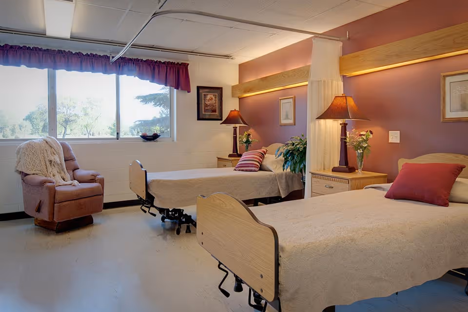 Two adjustable beds in a warmly decorated shared bedroom with bedside tables, lamps, a recliner by the window, and decorative pillows.