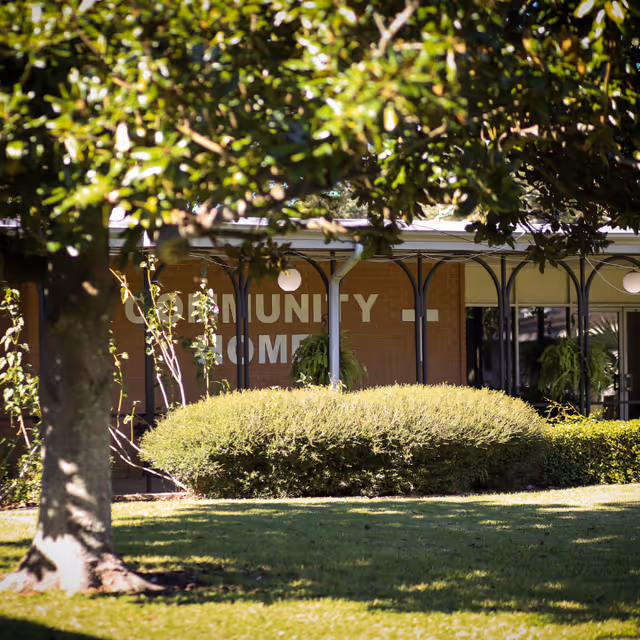 Front exterior of a community retirement home building partially obscured by trees and shrubs.