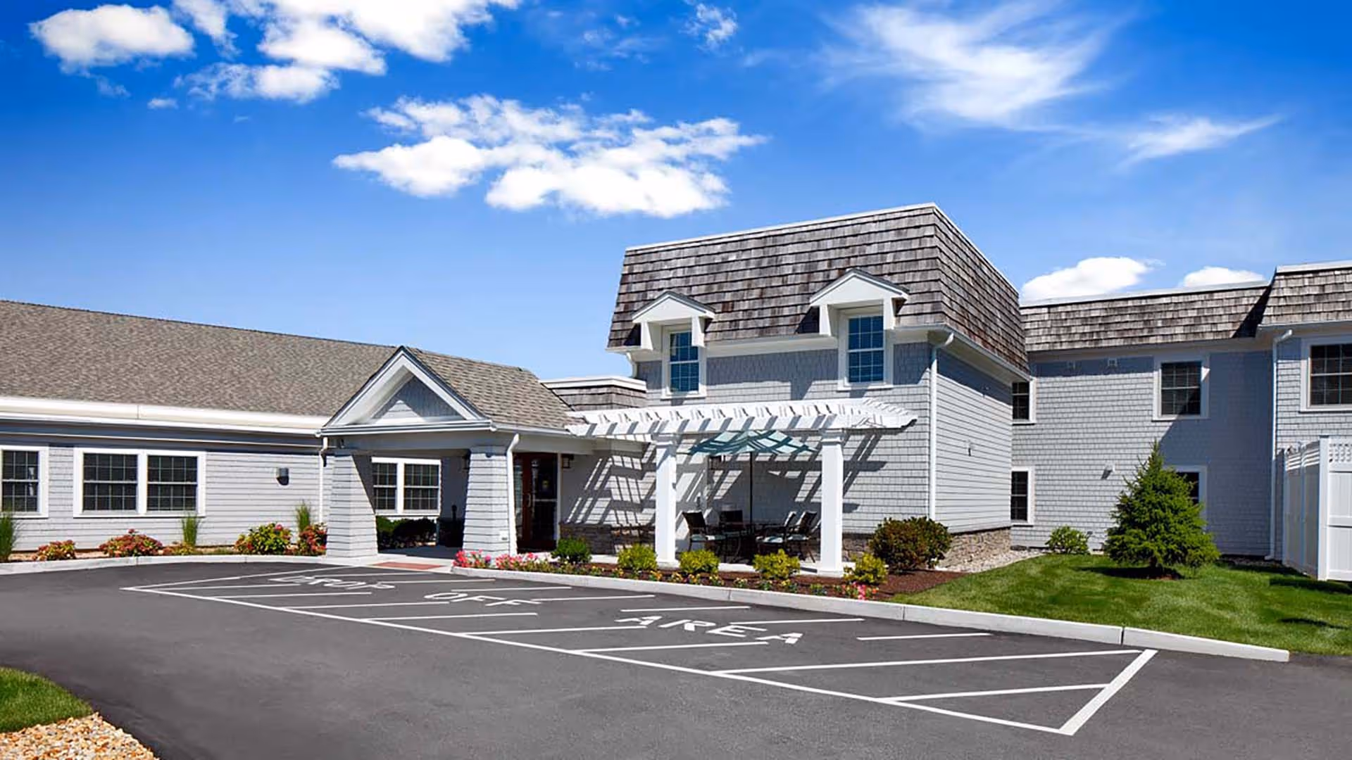 Front exterior of a senior living building with a covered entrance, pergola seating area, landscaped beds, and marked parking spaces under a blue sky.