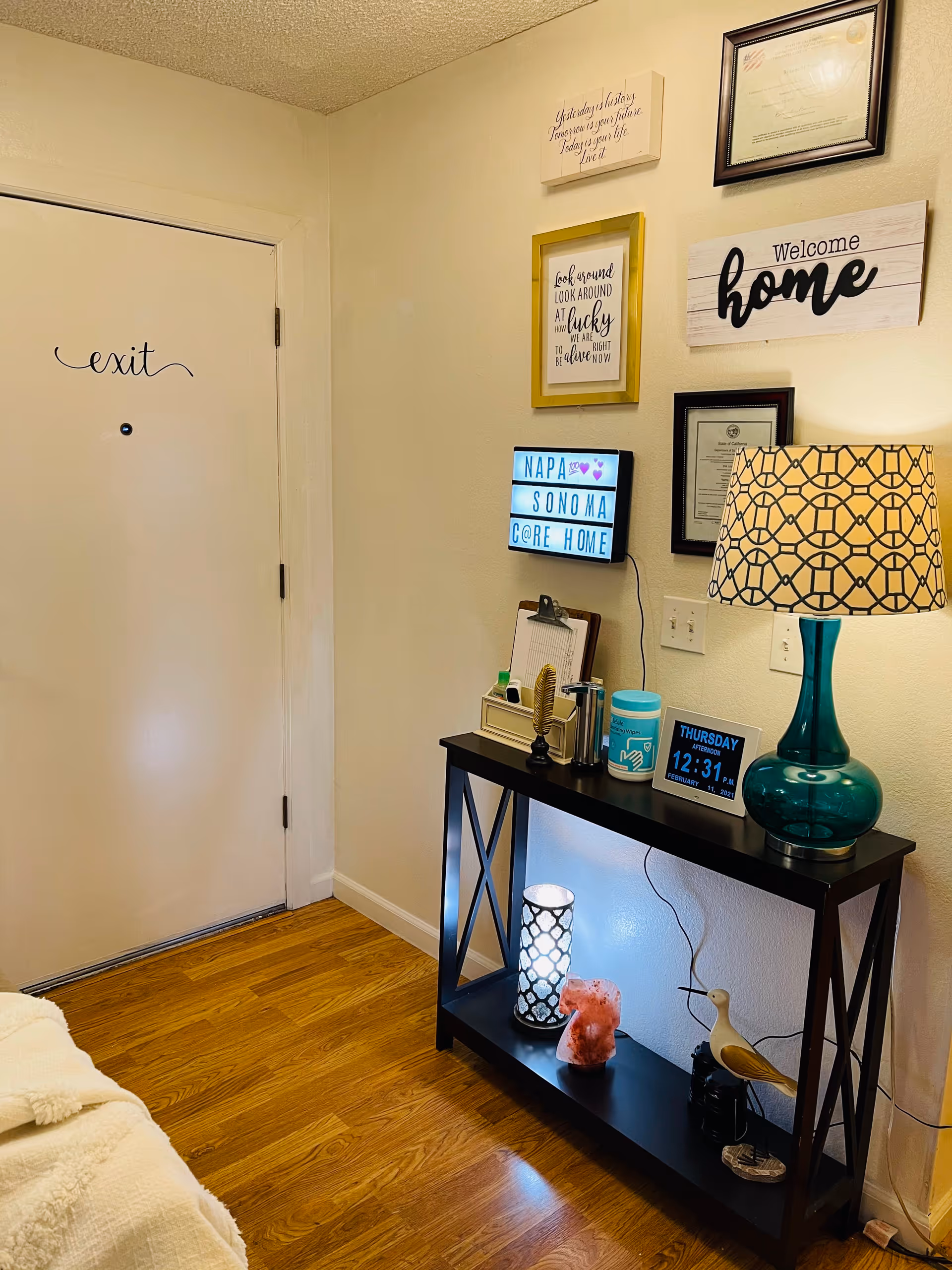 A cozy corner of a room with a white door labeled 'exit' on the left. A black console table with decorative items including a teal lamp with a patterned shade, a digital clock showing Thursday 12:31 PM, a small lightbox sign reading 'NAPA SONOMA CARE HOME', and various small decor pieces. Above the table are framed quotes and certificates, including a sign that says 'Welcome home'. The floor is wooden and the walls are light-colored.