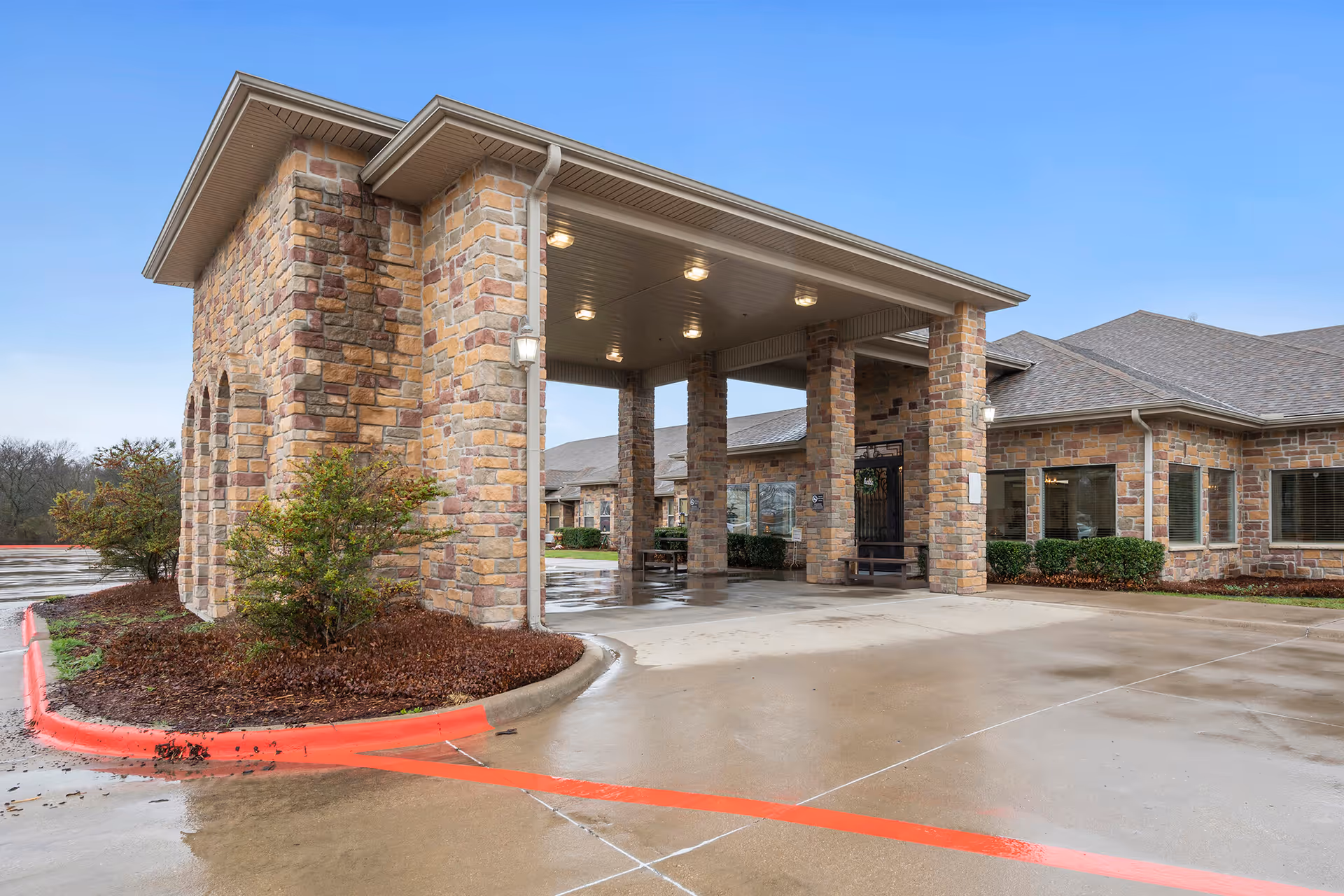 Entrance of a nursing and rehabilitation facility with a covered drop-off area supported by stone pillars, surrounded by a wet driveway and landscaped bushes under a clear sky.