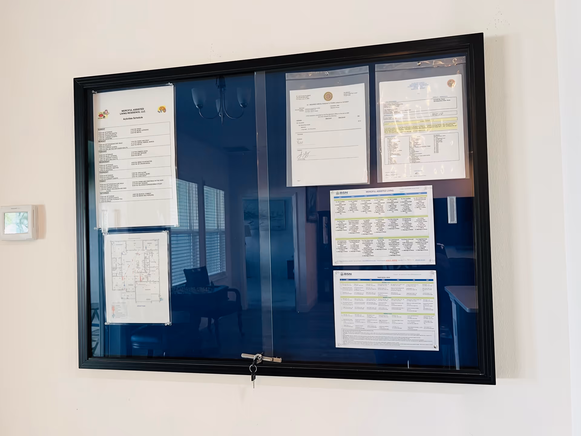 A black-framed bulletin board mounted on a white wall displaying various documents including an activity schedule, a floor plan, and other notices. The glass door of the board is locked with a key. Reflections of a room with chairs and windows are visible on the glass.