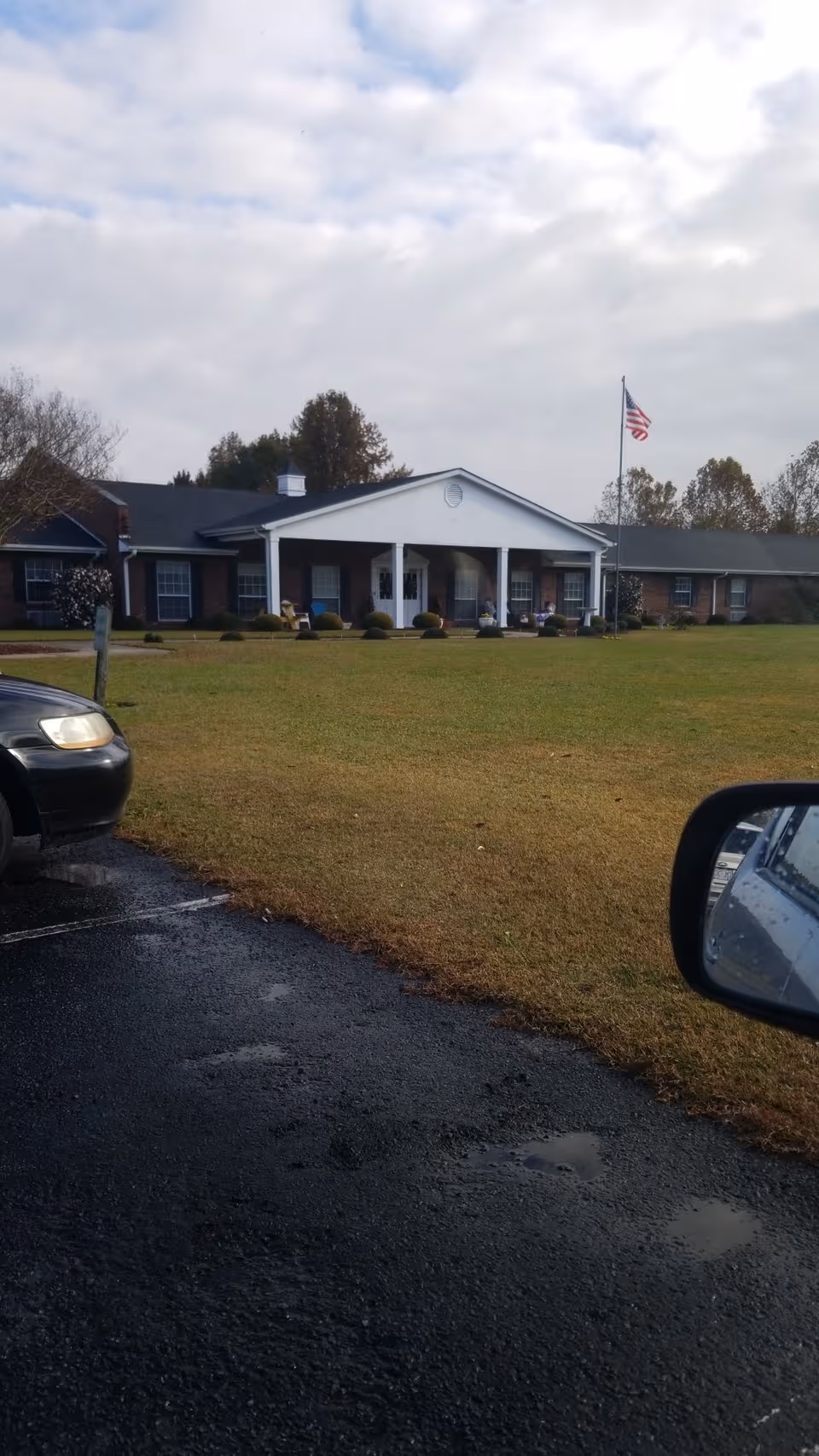 Assisted living facility exterior with white front porch, flagpole, and expansive lawn.