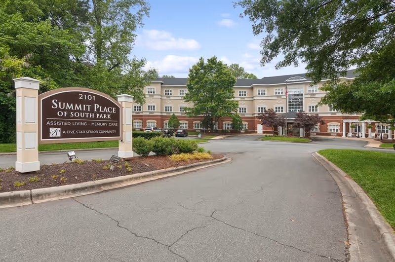 Exterior view of Summit Place of South Park, a senior living facility with a large sign in the foreground displaying the facility name and services. The building is three stories tall with a beige and brick facade, surrounded by trees and landscaping under a partly cloudy sky.