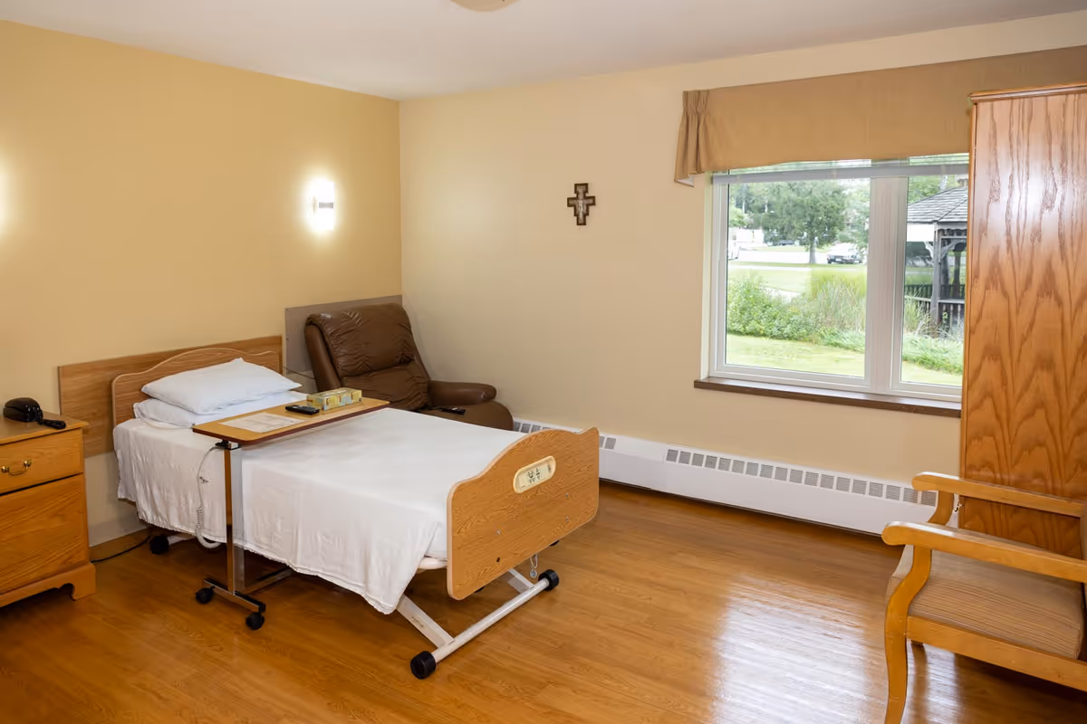 A single resident room with a hospital-style bed, recliner, dresser, and window view.