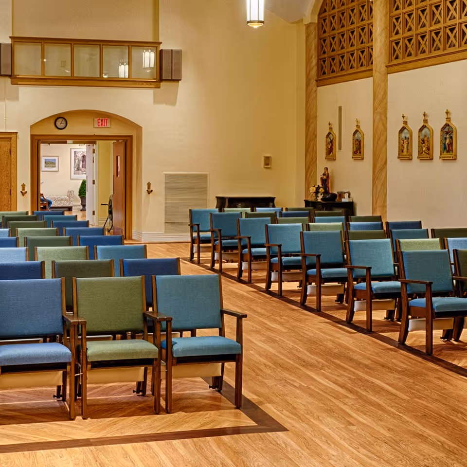 A bright interior room with rows of blue and green upholstered chairs on a wooden floor, religious plaques on the wall, and an exit doorway in the back.
