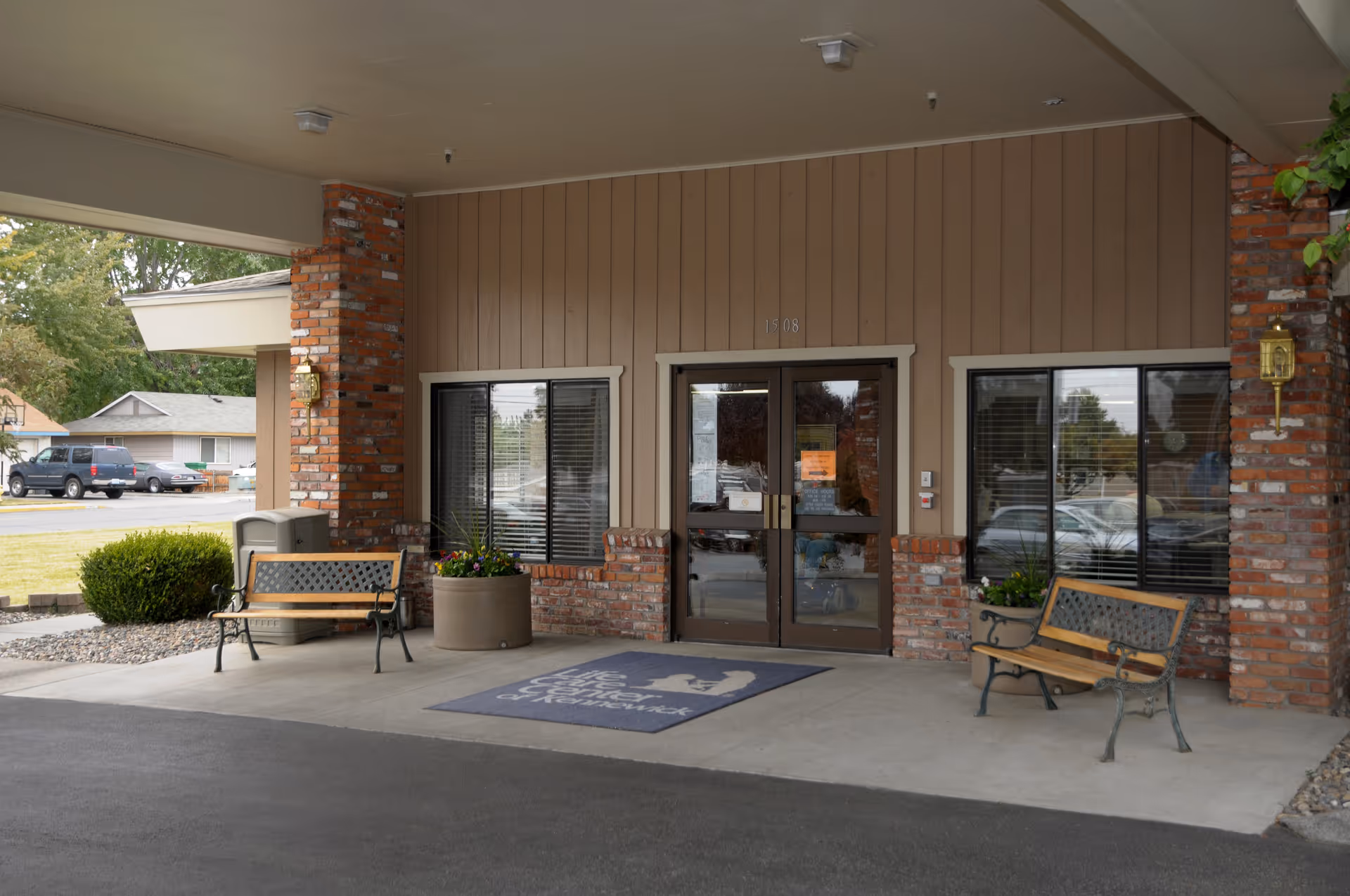 Covered front entrance of a brick-and-wood senior living facility with double glass doors, benches, planters, and a welcome mat.