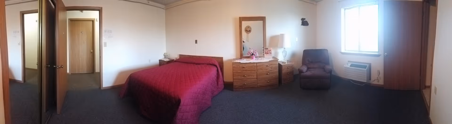 Panoramic view of a furnished senior living bedroom with bed, dresser, and chair.