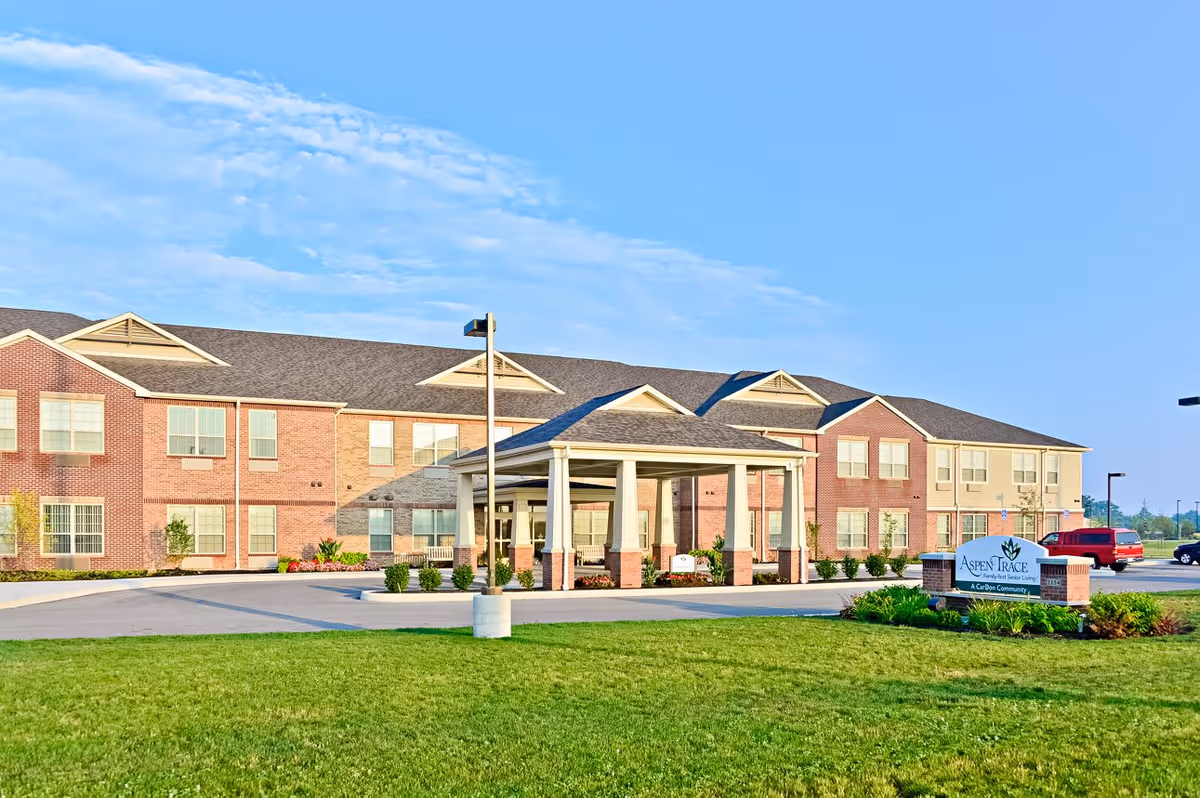 Exterior view of a two-story senior living facility named Aspen Trace with a covered entrance, well-maintained lawn, and a sign displaying the facility's name. The sky is clear and blue.