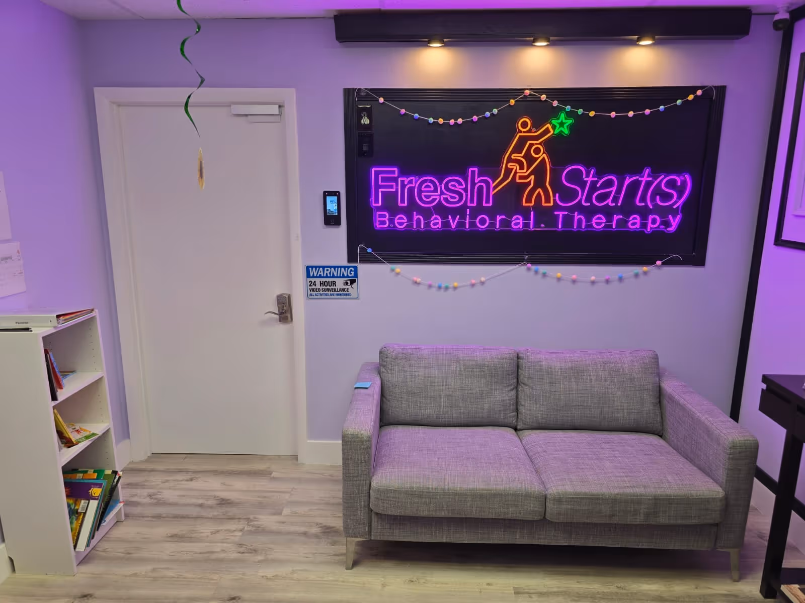 Lavender-walled therapy room with a neon 'Fresh Starts' sign and gray sofa.