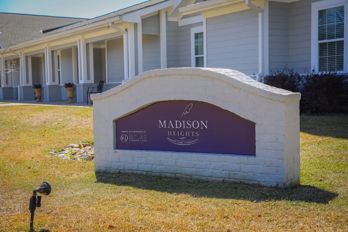 A white brick sign with a purple inset that reads 'MADISON HEIGHTS MEMORY CARE COMMUNITY' in front of a light gray building with white trim and windows. The sign also includes the logo and text 'OWNED AND OPERATED BY ATLAS SENIOR LIVING'. The area around the sign has grass and a small spotlight.