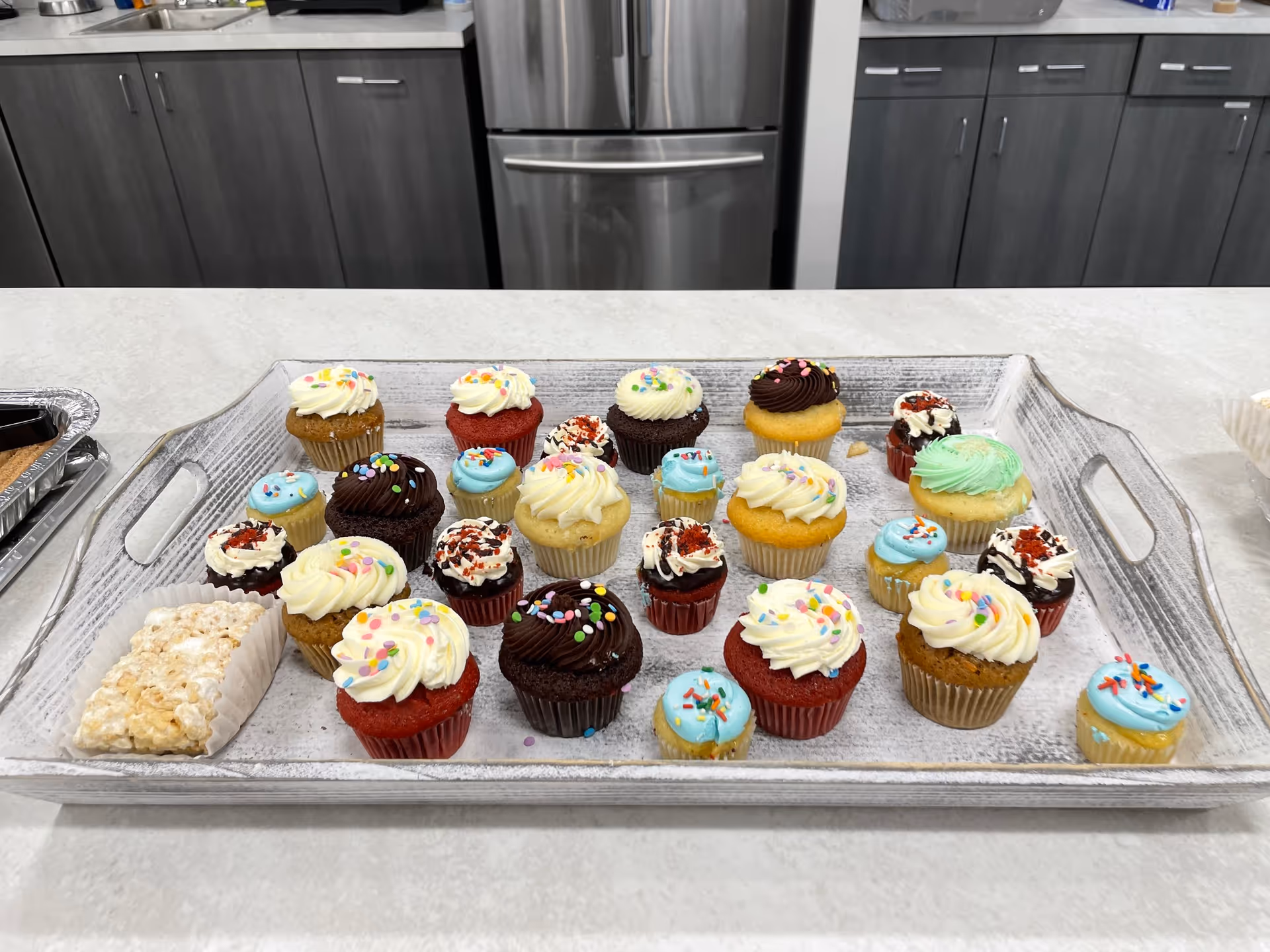 Tray of colorful cupcakes on a kitchen counter with a stainless steel fridge in the background.