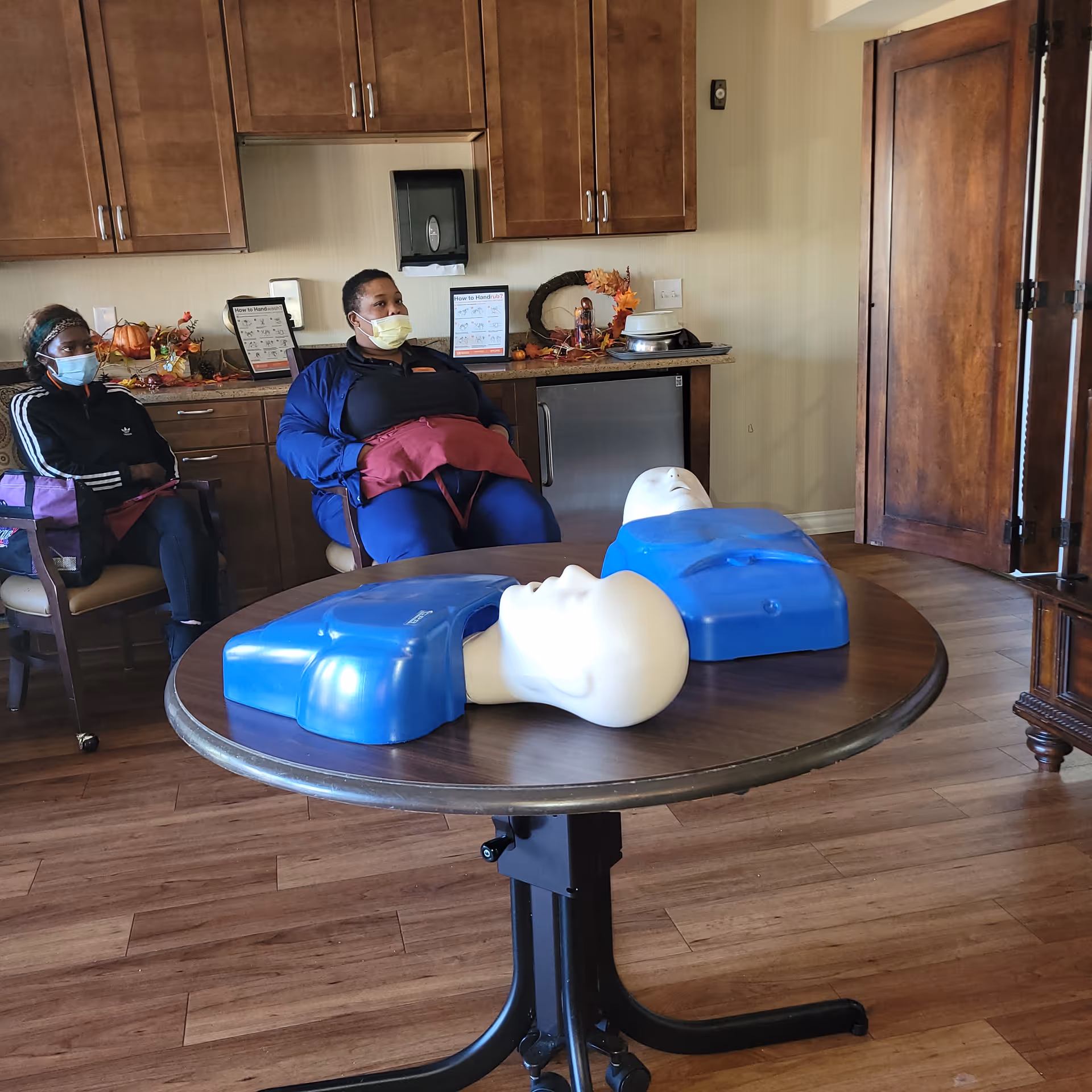 Two masked individuals sit in a training room with CPR mannequins on a round table.