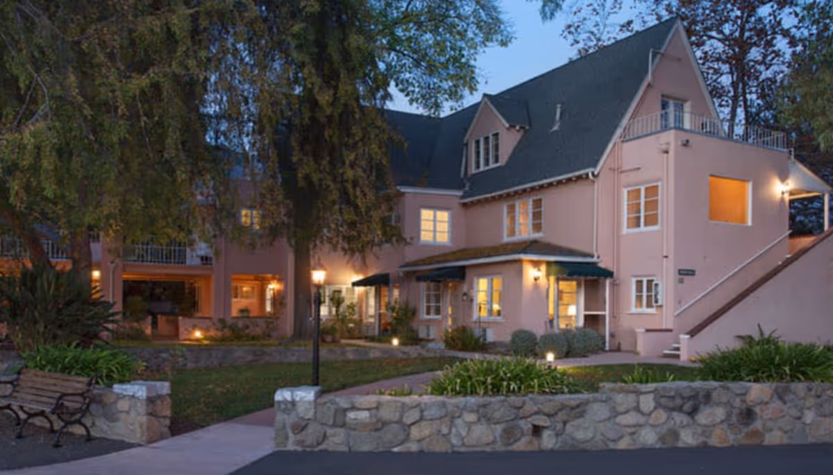 Pink, multi-story senior living building with illuminated windows, lawn, and stone wall facade.