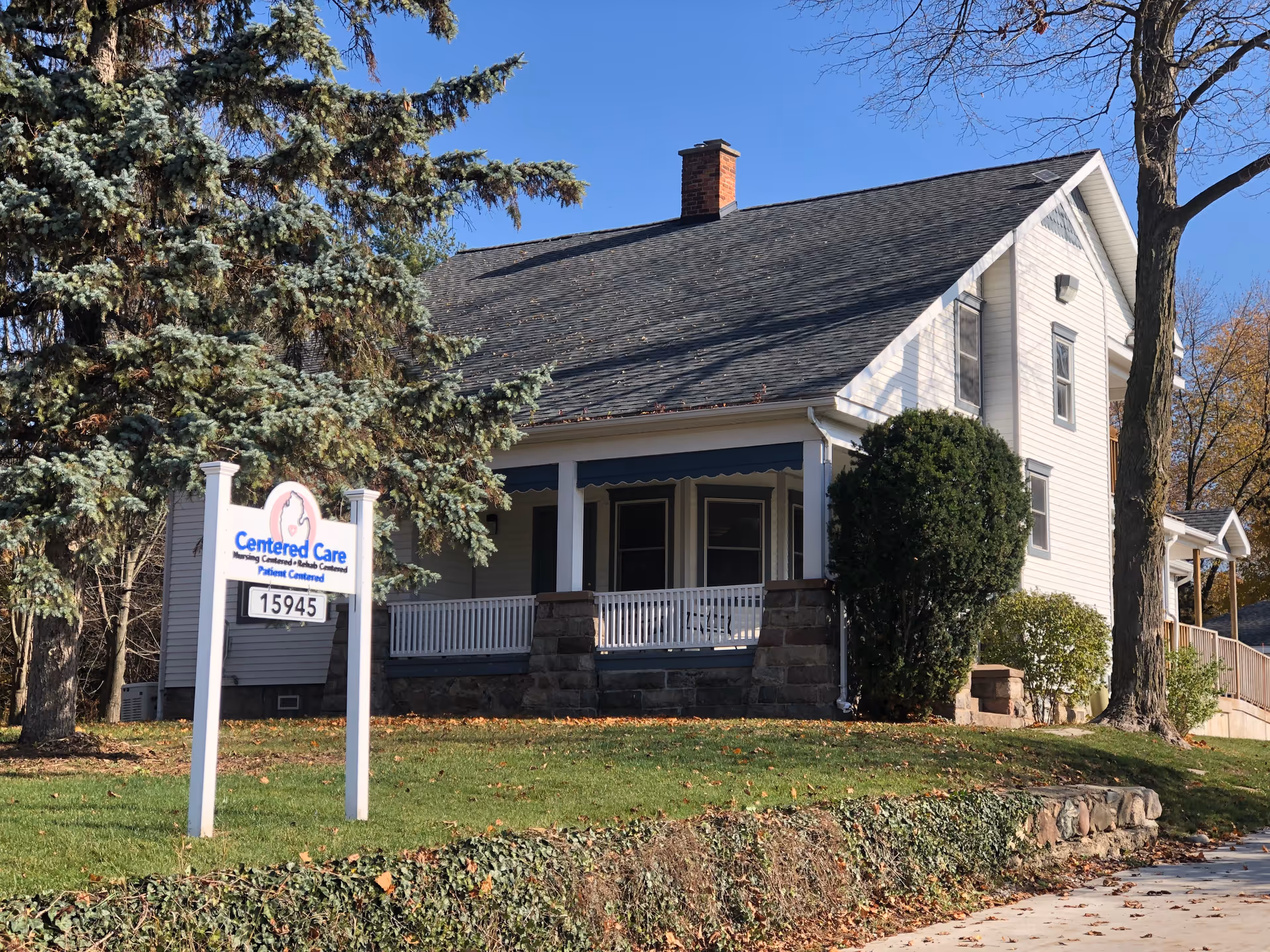 Exterior view of a senior care facility with sign, porch, and surrounding trees.