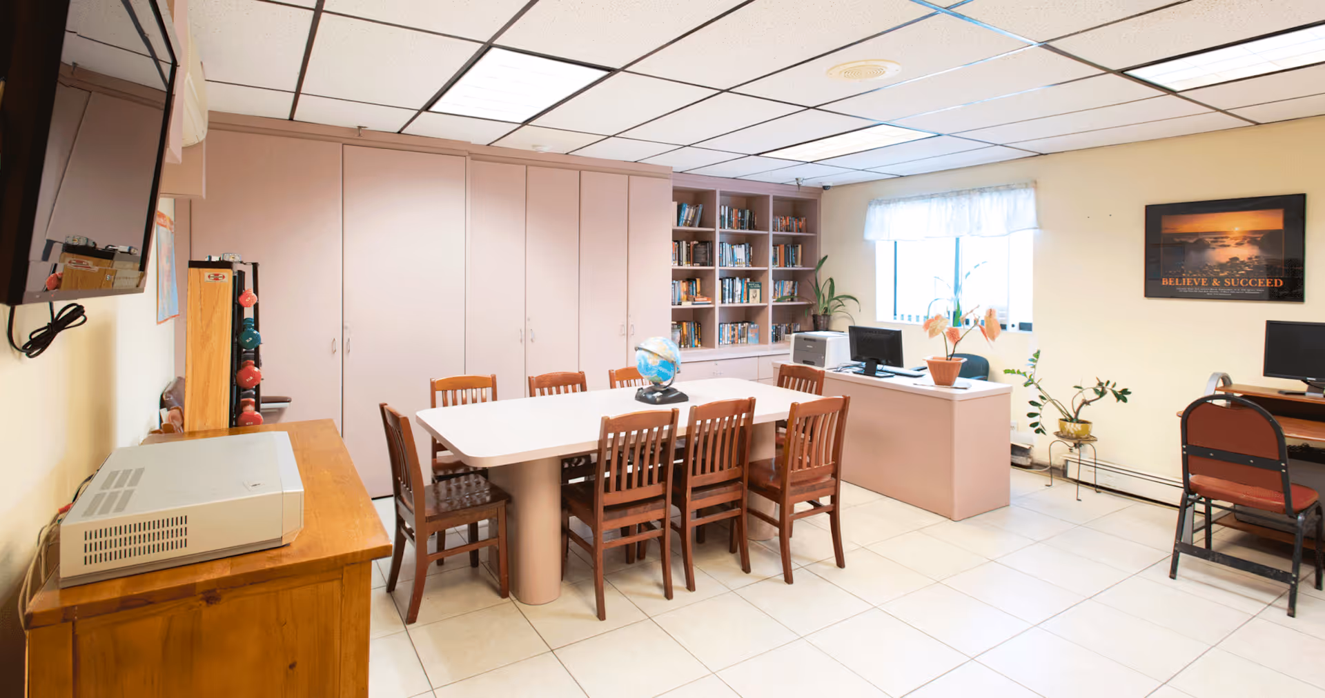 A well-lit room with a large rectangular table surrounded by wooden chairs. On the table is a globe. The room has built-in shelves filled with books, a desk with a computer, and several potted plants near a window with a white curtain. A motivational poster reading 'BELIEVE & SUCCEED' is on the wall. There is a television mounted on the wall and a wooden cabinet with a device on top.