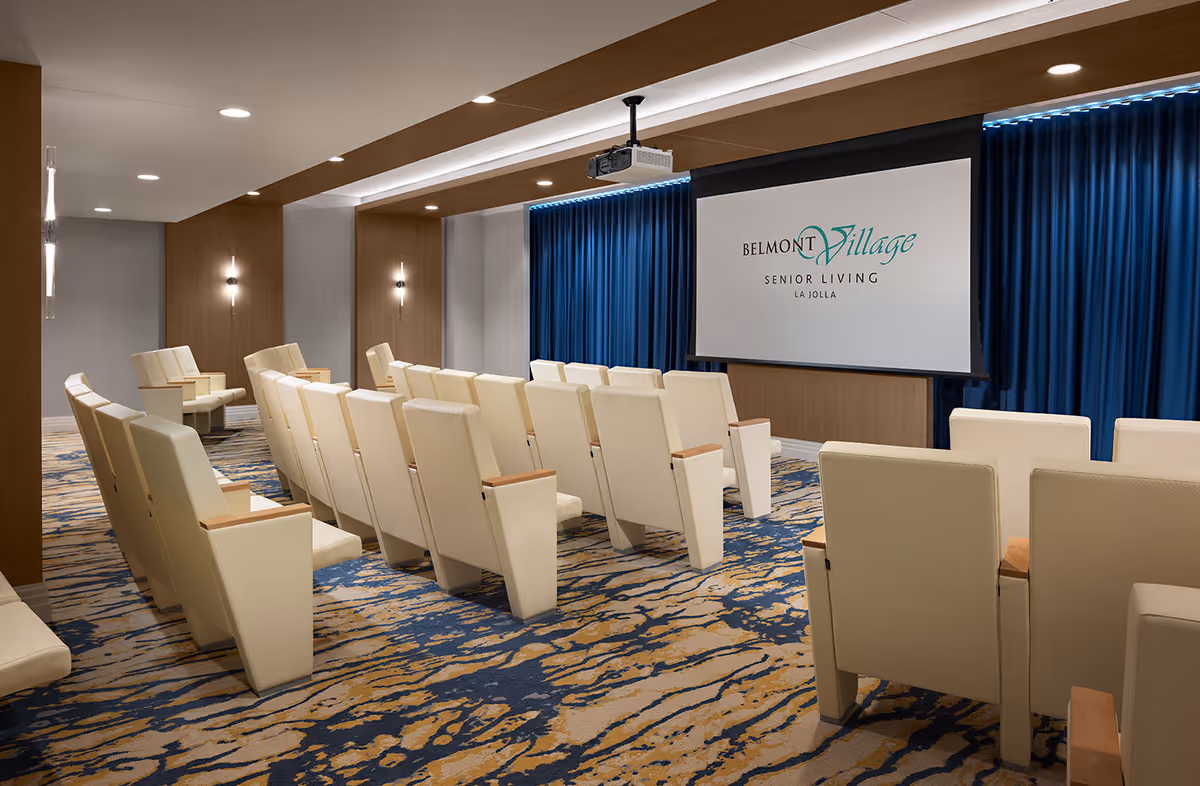 A modern presentation room with rows of beige cushioned chairs facing a large screen displaying the Belmont Village Senior Living La Jolla logo. The room features a patterned carpet in blue and beige tones, wood-paneled walls, recessed lighting, and a ceiling-mounted projector.