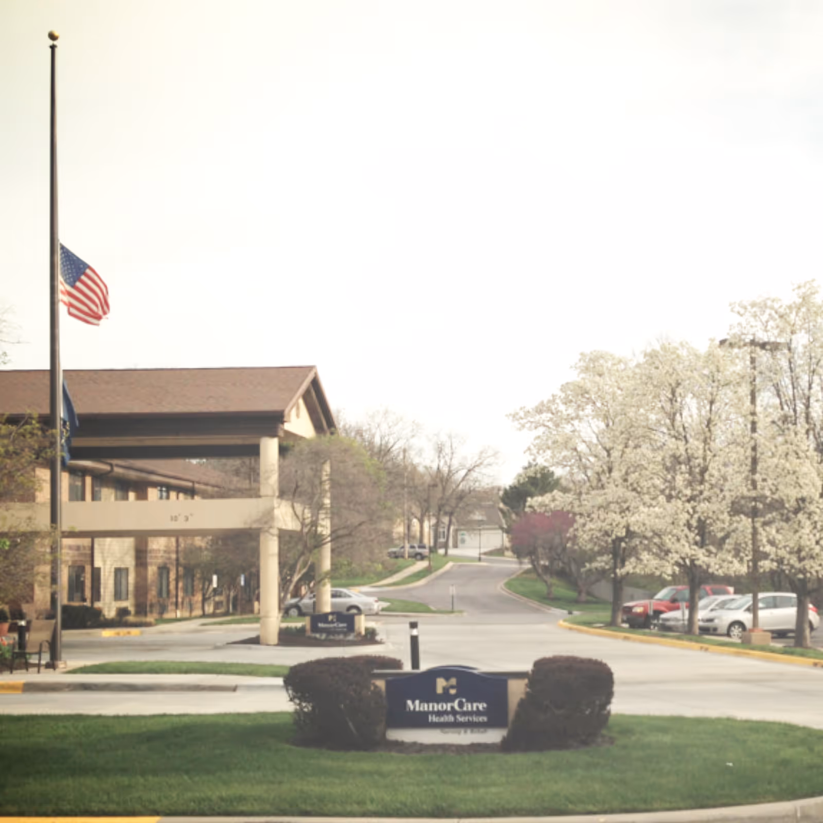 Exterior view of a senior living facility entrance with flag, sign, and manicured landscaping.