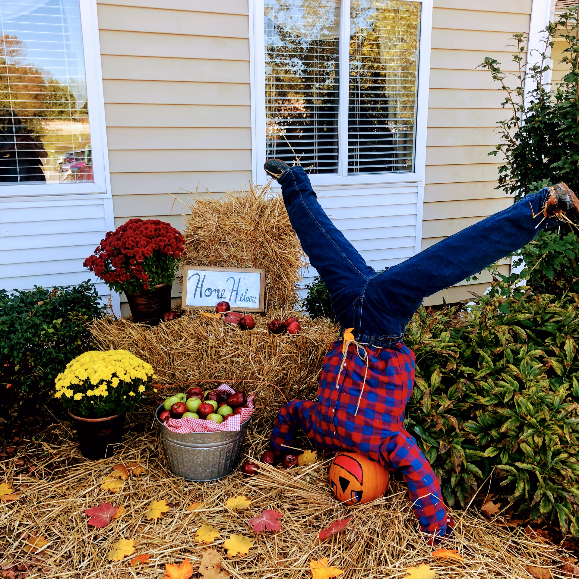 Fall display with hay bales, pumpkins, apples, and a scarecrow-like figure outside a house