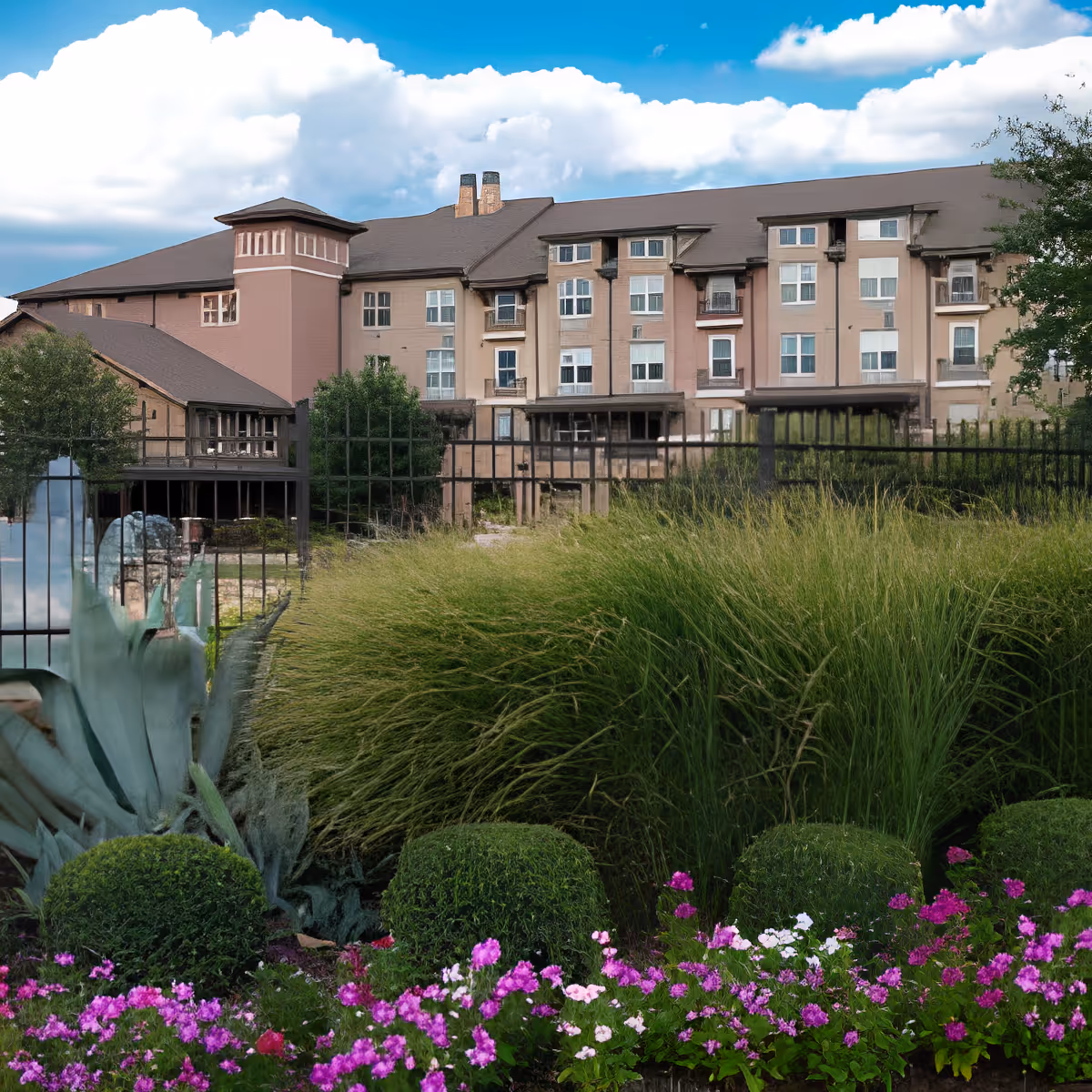 Exterior view of a multi-story senior living facility building with beige and pink walls, multiple windows, and a brown roof. In the foreground, there is a garden with green bushes, tall grasses, and colorful flowers under a partly cloudy blue sky.