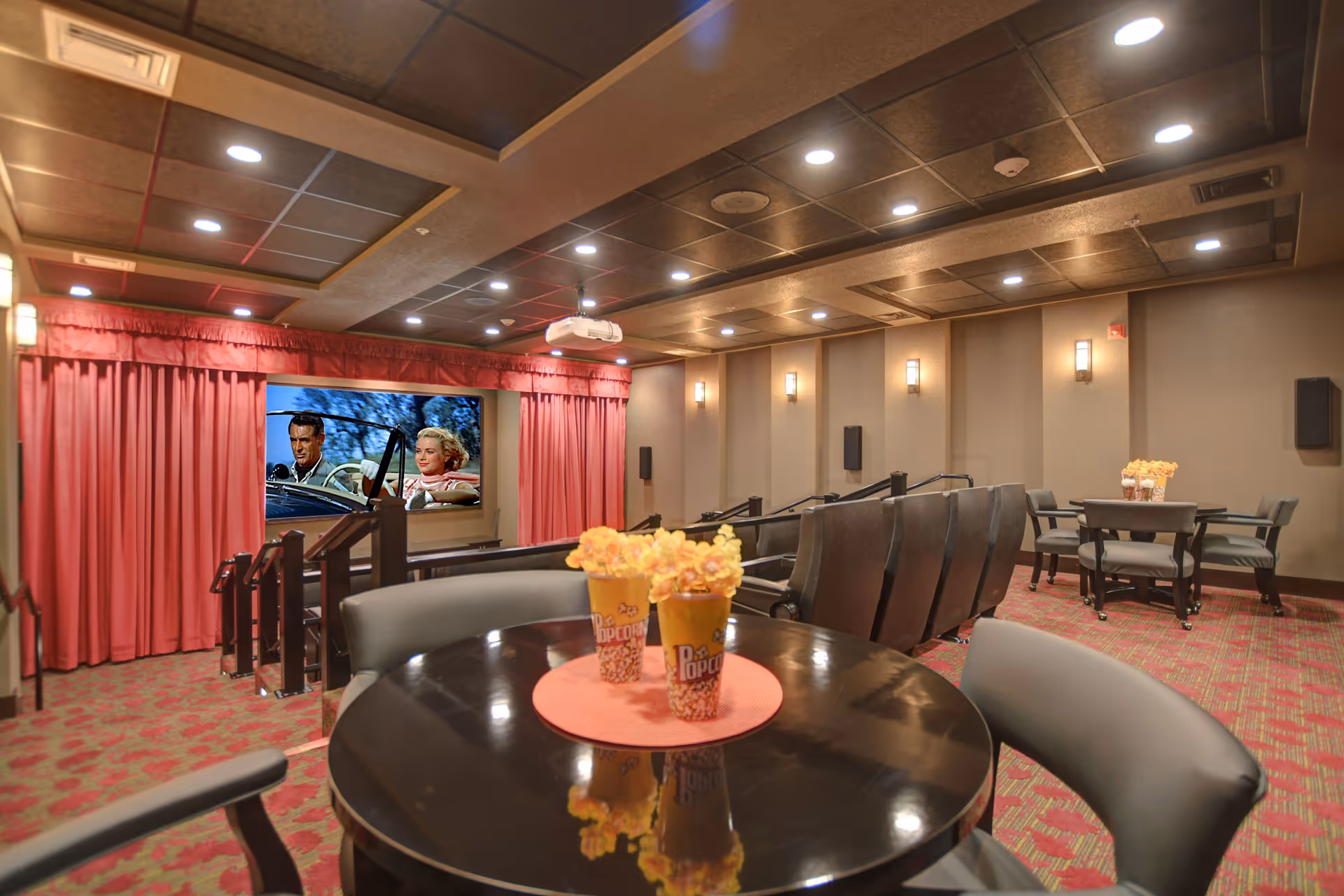 A cozy movie theater room with red curtains framing a large screen showing a classic film. The room has rows of comfortable theater seats and round tables with chairs in the foreground. Two popcorn containers with flowers are placed on the nearest table. The ceiling has recessed lighting and a projector is mounted above.