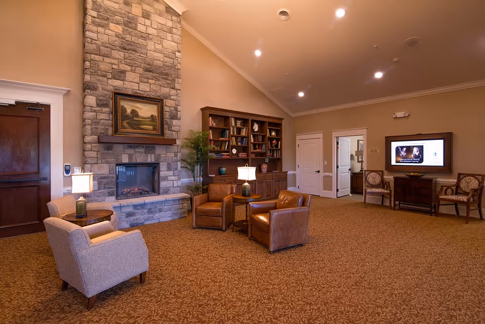 Spacious communal lounge with a stone fireplace, upholstered and leather chairs, a built-in bookshelf, and a wall-mounted TV.