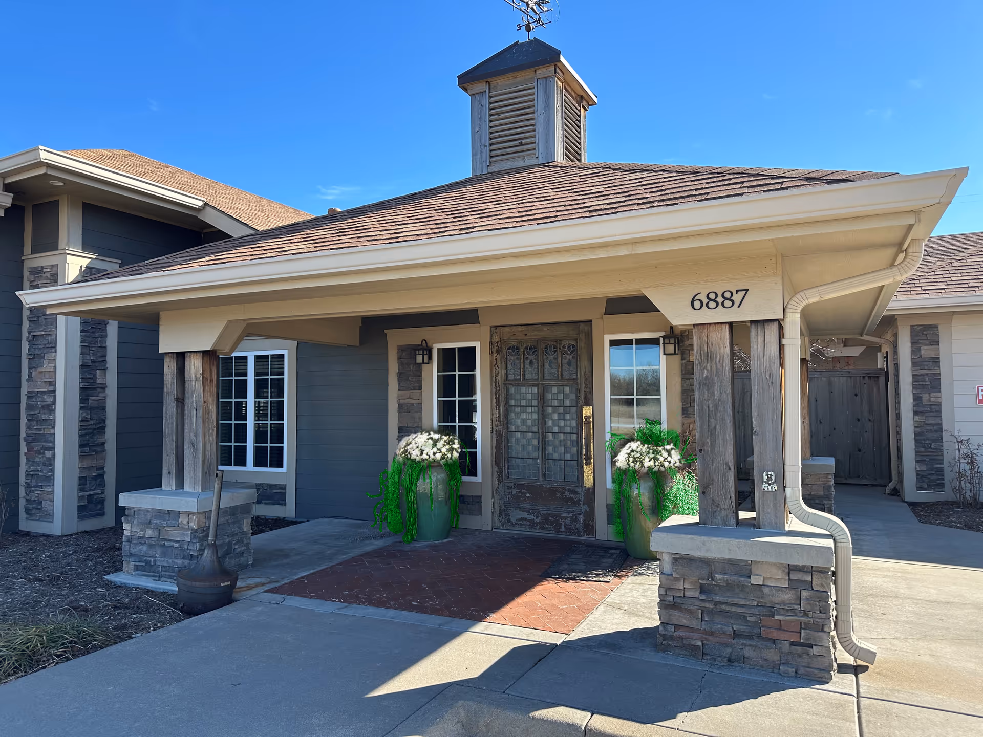 Front entrance of a senior living facility with a covered porch supported by wooden pillars and stone bases. The building has a shingled roof with a small cupola on top. Two large green planters with white flowers flank the wooden front door. The building number 6887 is displayed on the porch beam. The sky is clear and blue.