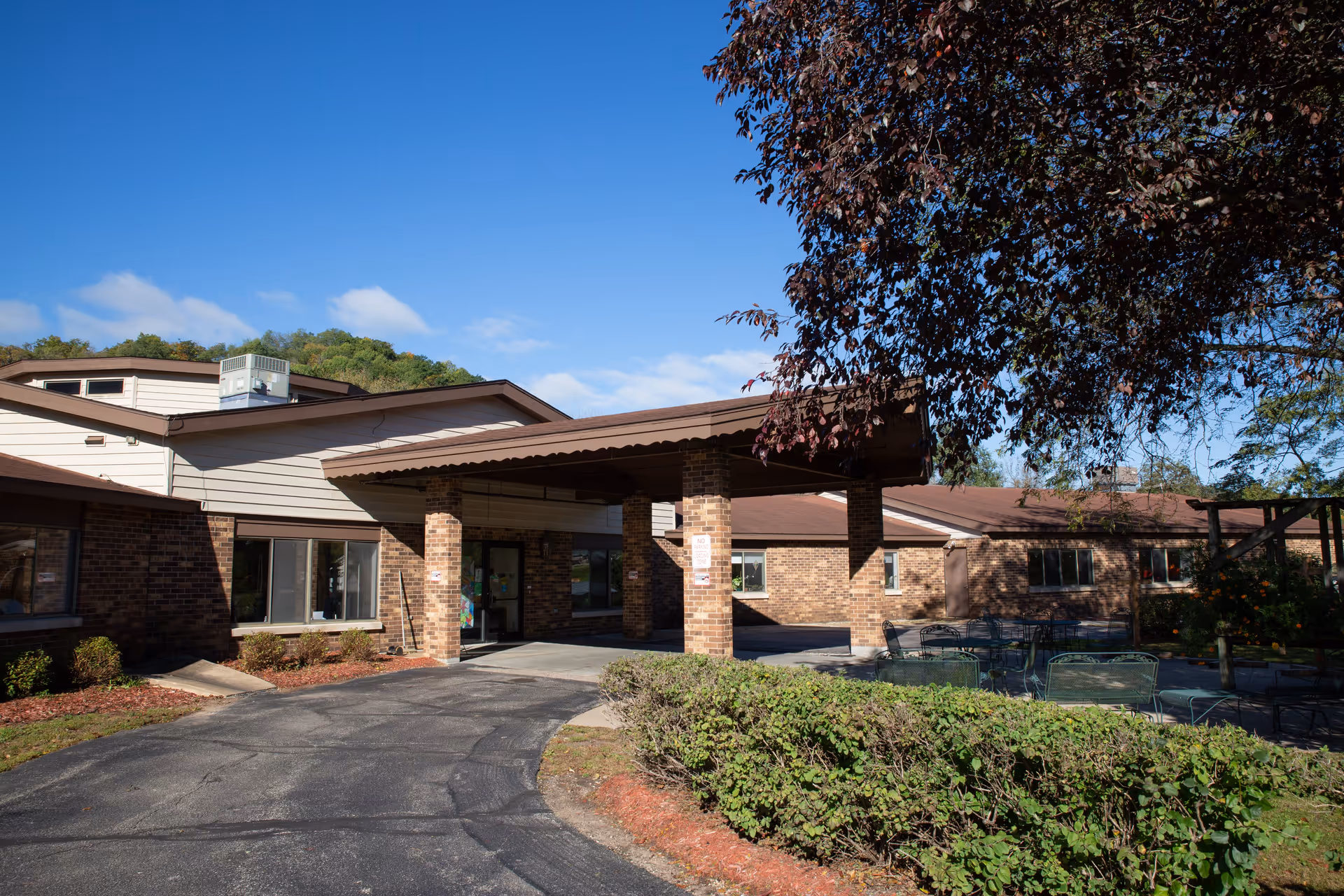Exterior view of Soldiers Grove Health Services building with a covered entrance, brick walls, and surrounding greenery including bushes and trees under a clear blue sky.