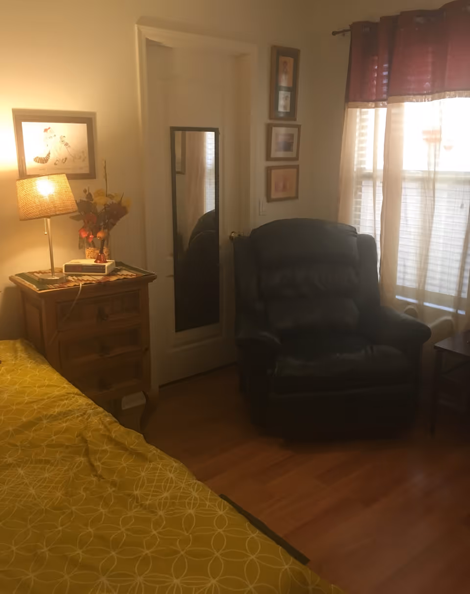 Cozy bedroom with yellow patterned bedspread, recliner, nightstand, mirror, and window.