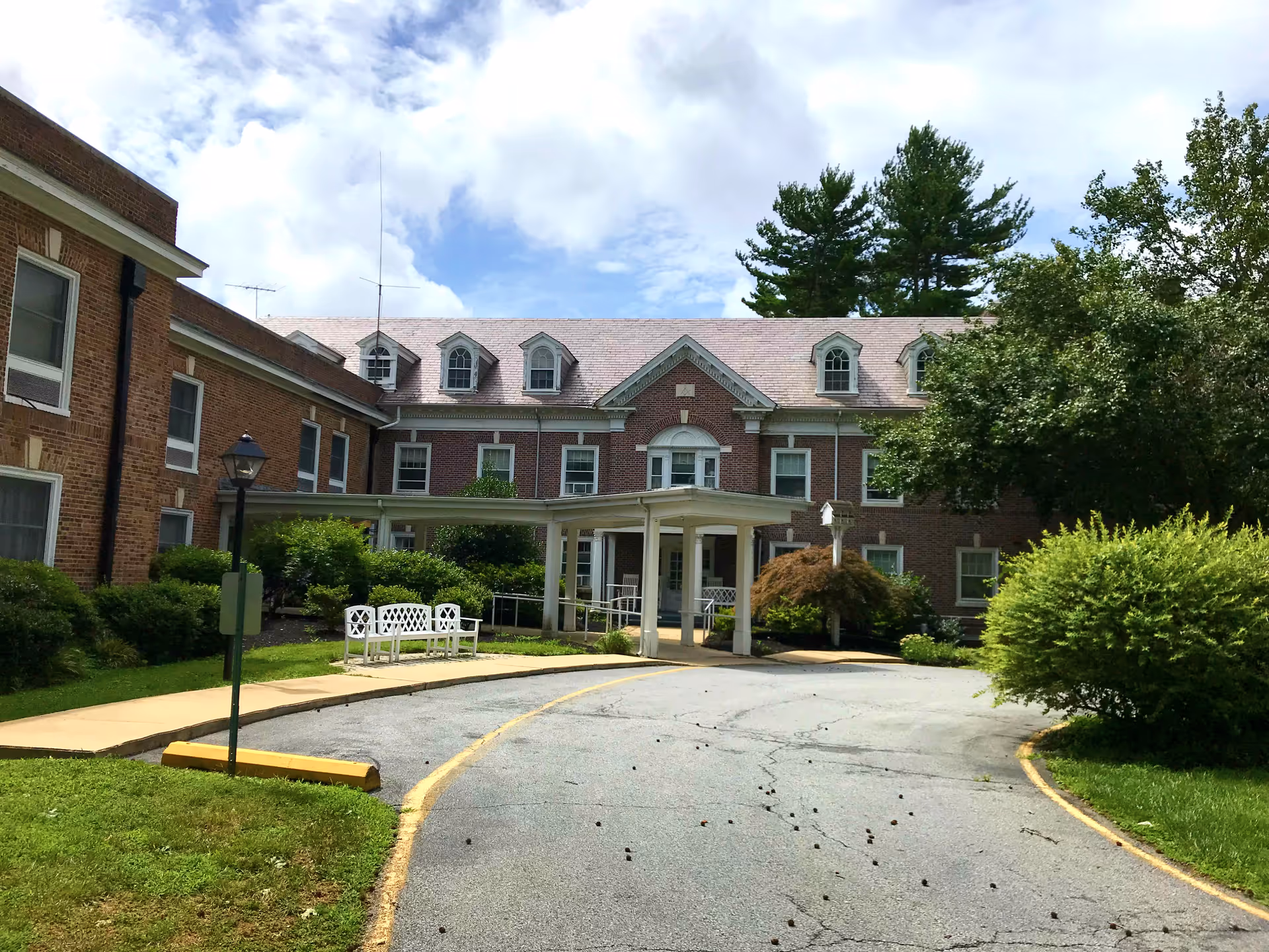 Brick senior living center with a covered entrance, curved driveway, and surrounding greenery.