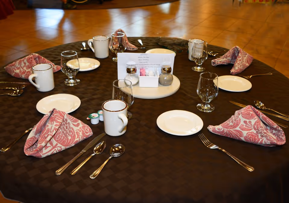 A round dining table set for six people with white plates, silverware, glassware, coffee mugs, and pink patterned cloth napkins folded neatly. In the center of the table is a rotating tray holding salt and pepper shakers, sugar packets, and a small sign. The tablecloth is dark brown and the floor is tiled.
