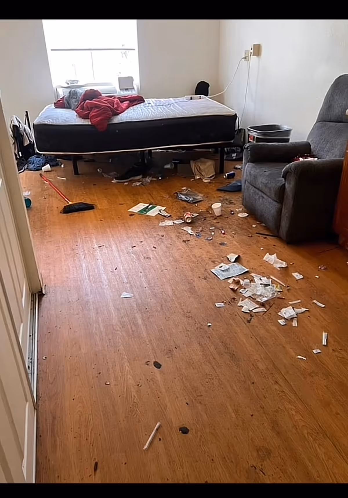 Messy bedroom with an elevated bed, chair, and scattered floor clutter in a senior facility.