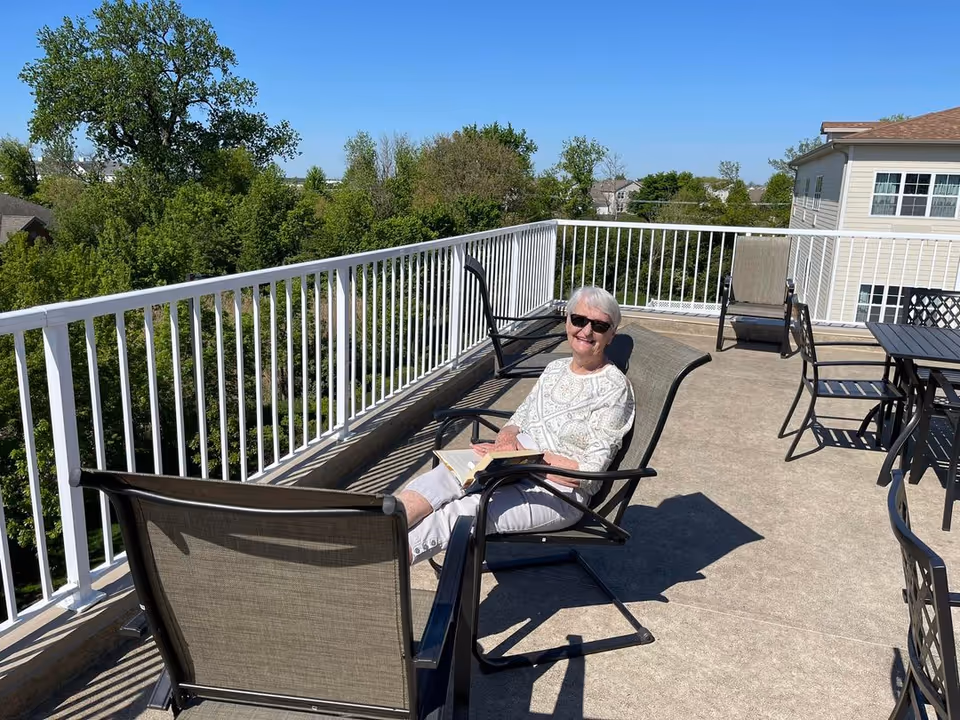 A smiling older woman wearing sunglasses relaxes in a lounge chair on a sunny rooftop patio with outdoor tables and trees beyond the railing.