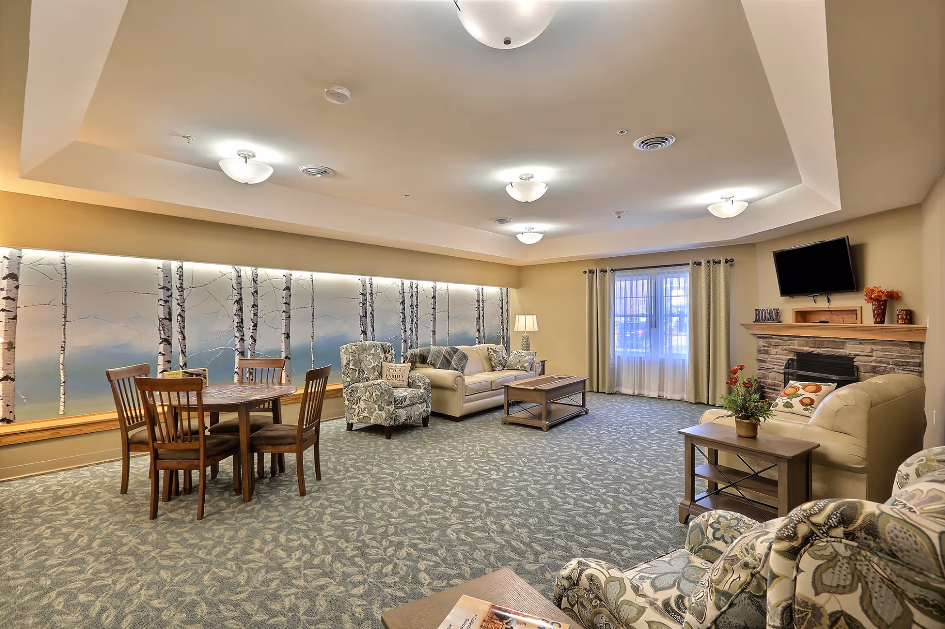A spacious senior living common area with patterned carpet, a wooden dining table with four chairs, two floral armchairs, a beige sofa, a coffee table, and a side table with a plant. The room features a wall mural of birch trees, a stone fireplace with a mounted TV above it, and a window with curtains letting in natural light.