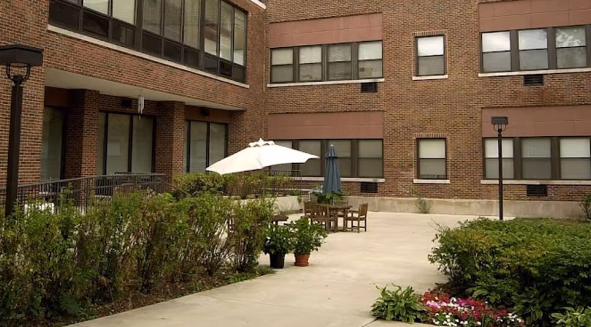 Brick building exterior with a paved courtyard, outdoor seating, umbrellas, and plants.