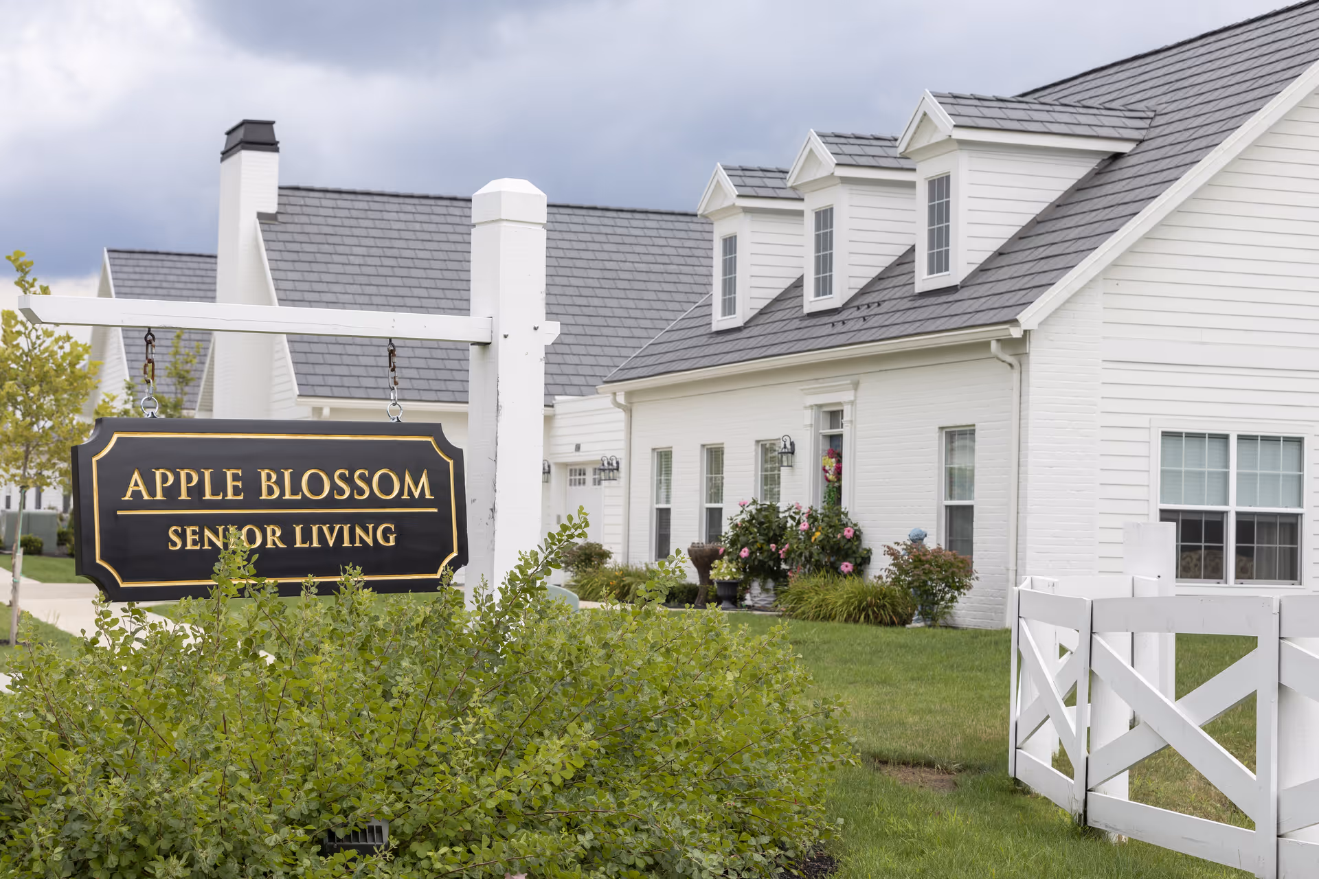 Exterior view of a white senior living facility building with a gray roof and three dormer windows. In the foreground, there is a black and gold sign that reads 'Apple Blossom Senior Living' hanging from a white post. The building is surrounded by green grass, bushes, and some flowers near the entrance.
