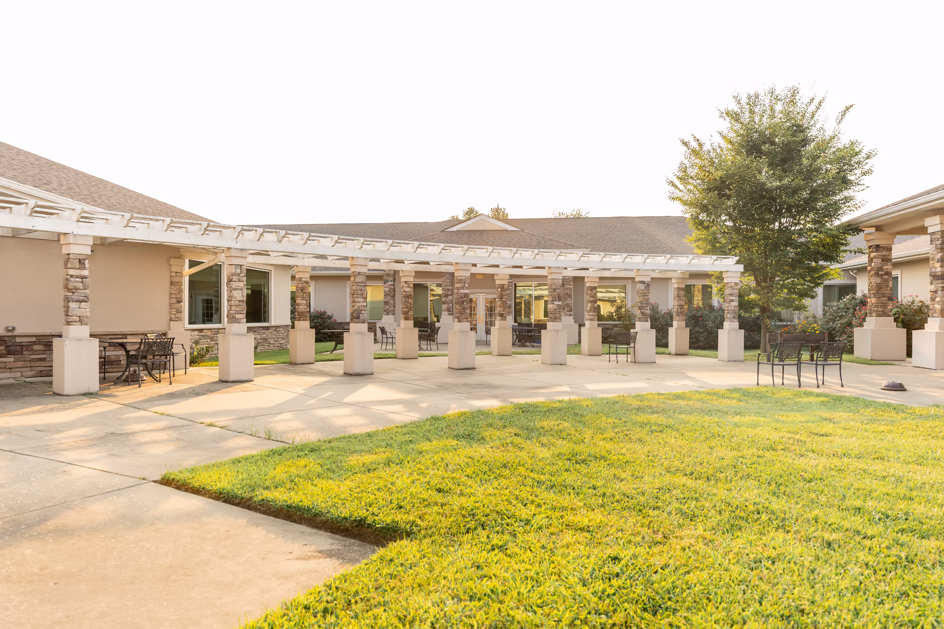 Outdoor courtyard area at Autumn Lake Healthcare at Waugh Chapel with a pergola supported by stone pillars, concrete walkways, green grass, several metal tables and chairs, and a tree on the right side.