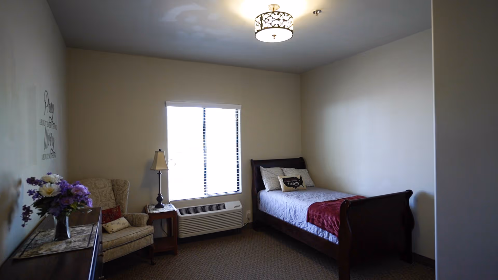 A furnished bedroom in a senior living facility with a bed, chair, and window.