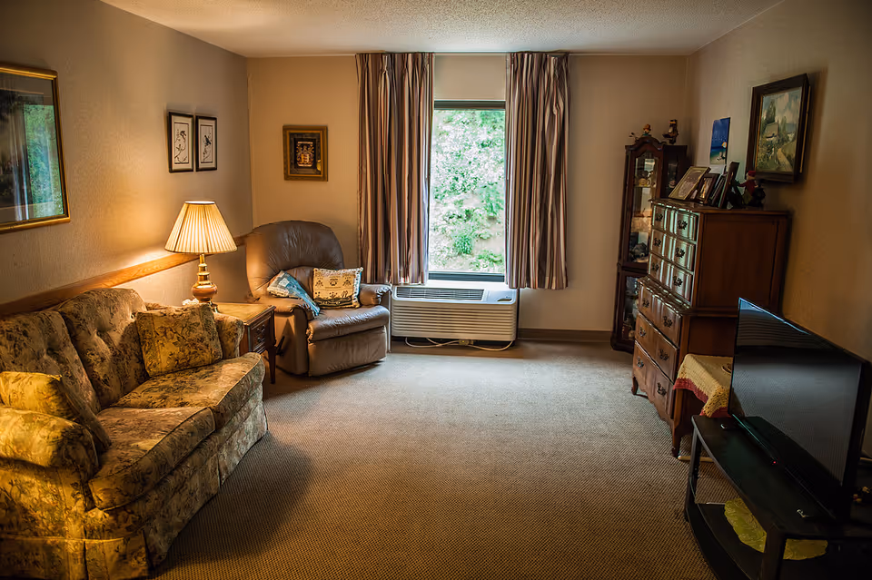 Cozy living room with a floral sofa, recliner and lamp, dresser and TV, centered around a window with an air conditioner.